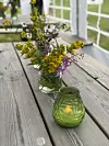 Blomster i glas og eit grønt lysglas på eit trebord ute.