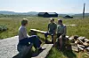 Tre personar sit ute ved ein grusveg med åkrar og fjell i bakgrunnen.