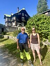 To personar står på ein grusveg framfor eit stort trehus med grøne tak.