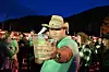 Mann i cowboyhatt held eit glas fram mot kameraet blant folk utandørs.