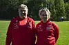 To personar i raude treningstrøyer står på ei grasbane.