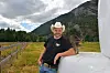 Mann i cowboyhatt står ved ei ku på eit beite framfor fjell.