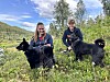 To personar med svarte hundar står i graset ved eit vatn.