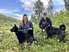 To personar med svarte hundar står i graset ved eit vatn.