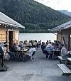 Mange gjester sit ved bord ute mellom to bygningar ved vatnet.