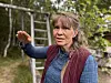 Kvinne gestikulerer framfor eit gjerde og ein stige i grøne omgjevnader.