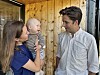 Kvinne held eit spedbarn medan ein mann står ved sida av dei framfor ein trevegg.