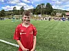 Gut i raud fotballdrakt står på ei grasbane med fotballaktivitet i bakgrunnen.