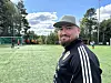 Skjegga mann i svart sportsdrakt står på ei fotballbane med spelarar bak seg.
