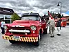 Raud veteranbrannbil står på eit torg med folk og andre brannbilar i bakgrunnen.