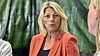 Blond kvinne i oransje jakke snakkar med to personar framfor ein grøn bakgrunn.
