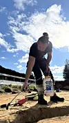 Arbeidar bøyer seg over ei grøft med handverktøy og ein behaldar i solskin.