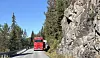 Raud buss køyrer på veg mellom ein bergvegg og barskog.