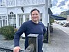 Mann står ved eit steingjerde framfor ein kvit trebygning, med fjell i bakgrunnen.