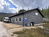 Grå trebygning ved ein grusveg, omgjeven av skog, med fjell og skyer i bakgrunnen.