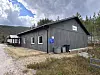 Grå trebygning ved ein grusveg, omgjeven av skog, med fjell og skyer i bakgrunnen.