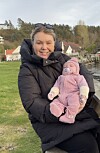 Kvinne i svart jakke held eit lite barn i rosa klede ved vatnet.