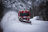 Raud brøytebil køyrer gjennom snø på ein skogveg.