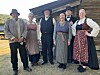 Seks personar i historiske klede står framfor ein trebygning.