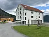 Lyst bygg med raudt tak ved eit grøntområde, med fjell og bustadhus i bakgrunnen.