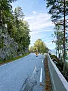 Ein person i oransje klede ferdast på ein skogkledd veg med fjell og fjord i bakgrunnen.
