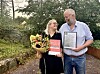 To personar står i ein hage med blomar og innramma dokument.