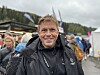 Smilande mann i svart jakke framfor ei folkemengd under open himmel.