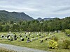Kyrkjegard med gravminne, tre og skogkledde fjell under overskya himmel.