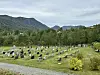 Kyrkjegard med gravminne, tre og skogkledde fjell under overskya himmel.