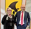 To personar står side om side framfor ein gul og svart logo innandørs.