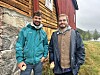 To menn står utanfor ein trebygning i eit grønt landskap.