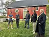 Fem personar står på graset utanfor ein raud trebygning.