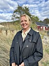 Kvinne i mørk frakk står i eit graslandskap framfor eit raudt hus.