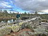 Person står på ein steinmura konstruksjon ved eit vatn i eit skogkledd fjellandskap.
