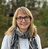 Kvinne med briller og skjerf poserer utandørs framfor grøne tre.