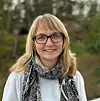 Kvinne med briller og skjerf poserer utandørs framfor grøne tre.