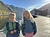 To barn står på ei trebru ved fjorden med fjell og bygning i bakgrunnen.