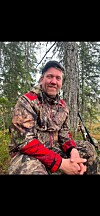 Mann i kamuflasjeklede sit framfor eit tre i skogen med eit jaktvåpen ved sida.