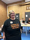 Kvinne i svart T-skjorte står i eit rom medan andre sit ved bord.
