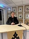 Kvinne sit ved eit bord med mikrofonar framfor ei vegg med innramma fotografi.