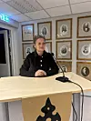 Kvinne sit ved eit bord med mikrofonar framfor ei vegg med innramma fotografi.