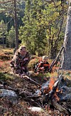 Ein vaksen og eit barn sit ved eit lite bål i skogen, med eit gevær lent mot eit tre.