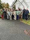 Gruppe vaksne står samla framfor ein kvit trebygning.
