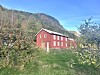 Raud trebygning i eit grønt landskap med blomstrande tre og skogkledde åsar.
