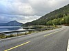 Asfaltert veg svingar langs ein fjord med skogkledde fjell og skyer.