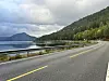 Asfaltert veg svingar langs ein fjord med skogkledde fjell og skyer.