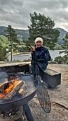 Person ved open eld i bålpanne, med vatn og fjell i bakgrunnen.