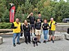 Gruppe menneske står ute med ein hund framfor tre og eit raudt banner.