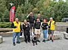 Gruppe menneske står ute med ein hund framfor tre og eit raudt banner.