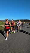 Løparar spring på ein asfaltveg i solskin, med åsar og jordbruksland i bakgrunnen.